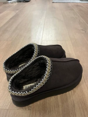 Bruna tofflor med fuskpäls - Mysiga bruna skor  med mjukt foder i fuskpäls och dekorativt mönster runt öppningen. Perfekta för att hålla fötterna varma