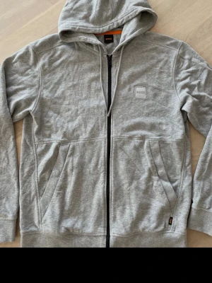 Grå zip-hoodie från BOSS - Snygg grå hoodie från BOSS med dragkedja framtill, huva med dragsko och två stora fickor. Diskret BOSS-logga på bröstet och mjukt material som känns skönt mot huden. Perfekt för en avslappnad och stilren look.