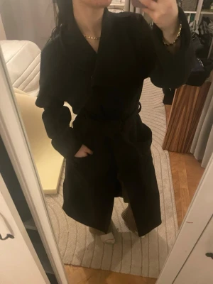 Svart kappa 50% Ull med bälte och bred krage - Stilren svart unik  ullkappa med bred krage och knytskärp i midjan. Kappan har en klassisk, rak passform och är perfekt för kyligare dagar. Fickor på sidorna och mjukt material som ger en snygg siluett. Enkel att matcha med olika outfits. Jag på bild är 157 och xs/s. Använd gärna köp nu