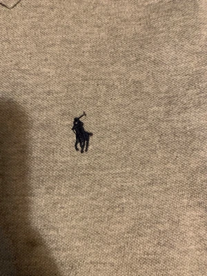 Grå pikétröja från Ralph Lauren - Klassisk grå pikétröja från Ralph Lauren med korta ärmar och krage. Tröjan har två knappar framtill och den ikoniska broderade loggan i marinblått på bröstet. Perfekt till jeans eller shorts för en clean och stilren look.