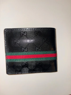 Svart plånbok  med ränder - Snygg svart plånbok präglad med GG-loggor och klassiska gröna och röda ränder på utsidan. Tillverkad i skinn med flera kortfack på insidan. Perfekt för dig som vill ha en stilren och lyxig accessoar.