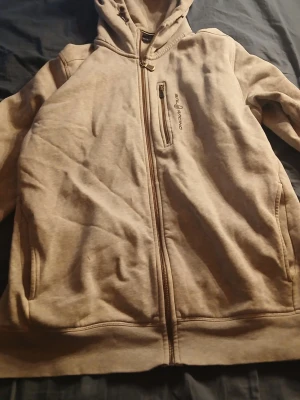 Grå hoodie från Sail Racing - Säljer en grå hoodie från Sail Racing med dragkedja framtill och en bröstficka med logga. Hoodien har huva med dragsko och två sidofickor. Insidan är mjuk och fleecefodrad för extra komfort. Perfekt för chill dagar eller när du vill ha något bekvämt.