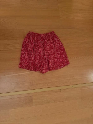 Röda blommiga shorts med resårmidja - Säljer ett par röda shorts med små blommor i gult, rosa och vitt. De har en bekväm resår i midjan och är tillverkade i ett lätt och luftigt tyg, perfekta för varma dagar. Passformen är loose och de har en avslappnad look.