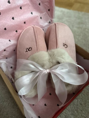 Rosa fluffiga tofflor med rosett - Supergulliga rosa tofflor från OX med mjukt fuskpälsfoder och stor satinrosett framtill. Perfekta för att hålla fötterna varma och mysiga hemma. Tofflorna har en platt sula och broderad logga på ovansidan.