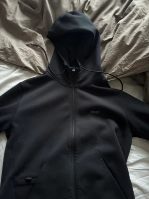 Sv soppsvart hoodie från BOSS - Säljer en svart hoodie från BOSS med dragkedja framtill och diskret BOSS-logga på bröstet. Hoodien har huva med dragsko och två fickor framtill. Perfekt för en clean och stilren look. Materialet känns mjukt och bekvämt.