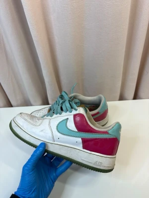 Nike Air Force 1 (Limited Edition 2007) - Nike Air Force 1 (Limited Edition 2007) sneakers med vit bas, turkosa detaljer och röd panel vid hälen. Skorna har klassisk snörning, rund tå och platt sula. Swoosh-loggan är turkos och materialet är en mix av skinn och syntet. Perfekt för dig som gillar färgstarka sneakers.