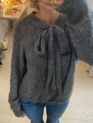 Mörkgrå stickad tröja med rosett - Mysig mörkgrå stickad tröja med lång ärm och en stor dekorativ rosett vid halsen. Tröjan har en fluffig, lite luddig struktur och avslappnad passform. Perfekt för kalla dagar och enkel att styla med jeans eller kjol.