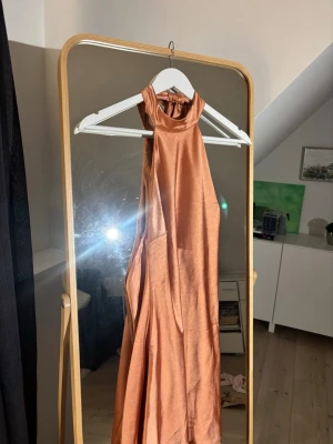 Persikofärgad halterneckklänning satin - Snygg persikofärgad kortklänning i satin med halterneck och öppen rygg. Klänningen har en lös passform och knyts i nacken med ett brett band. Perfekt för sommarens alla tillfällen.Klänningen har en liten fläck , men den syns knappt.