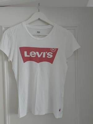 Vit Levi's t-shirt med röd logga - Klassisk vit t-shirt från Levi's med den ikoniska röda loggan tryckt framtill. T-shirten har rund halsringning och korta ärmar. Tillverkad i mjuk bomull för en skön och avslappnad look.