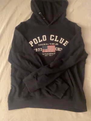 Polo Club hoodie - Mörkblå hoodie från Polo Club med vit text och amerikansk flagga på bröstet. Klassisk huva med dragsko och ribbade muddar vid ärmar och nederkant. Perfekt för en avslappnad och sportig stil.