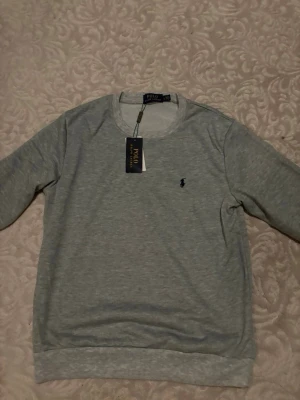 Grå sweatshirt från Polo Ralph Lauren - Klassisk grå sweatshirt från Polo Ralph Lauren med rund halsringning och den ikoniska mörkblå logotypen broderad på bröstet. Tröjan har ribbade muddar vid ärmslut och nederkant. Perfekt för en avslappnad och stilren look.