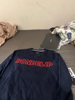 Mörkblå sweatshirt från Bondelid - Mörkblå sweatshirt från Bondelid med röd broderad logga över bröstet. Klassisk rund halsringning och ribbade muddar. Perfekt för en avslappnad och stilren look.