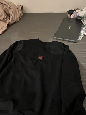 Svart sweatshirt från Hugo Boss - Svart sweatshirt från Hugo Boss med diskret broderad logga och en liten röd detalj framtill. Tröjan har rund halsringning och är tillverkad i mjukt material, perfekt för en stilren och avslappnad look.