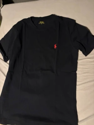 Svart t-shirt från Polo Ralph Lauren - Klassisk svart t-shirt från Polo Ralph Lauren med röd broderad logga på bröstet. T-shirten har rund halsringning och korta ärmar. Tillverkad i mjuk bomull för en skön känsla och enkel stil.