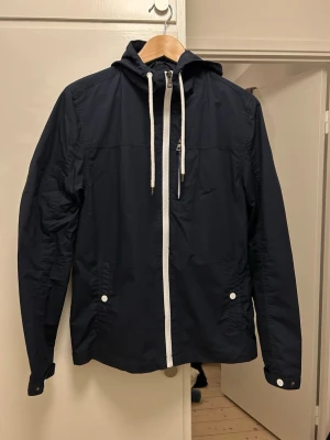 Mörkblå vindjacka från pier one - Snygg mörkblå vindjacka från pier one med vit dragkedja och vita snören i huvan. Jackan har två fickor med knappar framtill och justerbara ärmslut. Perfekt för blåsiga dagar och enkel att matcha med olika outfits.