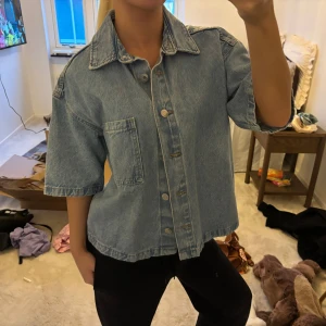 Ljusblå croppad jeansjacka - Trendig ljusblå jeansjacka med kort, boxig passform och korta ärmar. Jackan har klassisk krage, stora knappar framtill och en stor bröstficka. Perfekt för en avslappnad och cool stil.