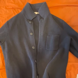 Mörkblå kofta från Kappahl - Stilren mörkblå kofta från Kappahl med knäppning framtill och en bröstficka. Koftan har klassisk krage och är tillverkad i mjukt material som passar perfekt för lager på lager. Enkel och snygg design som funkar till många olika looks.