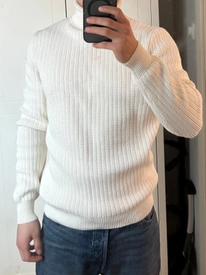 Vit ribbstickad polotröja - Säljer en stilren vit ribbstickad polotröja med hög krage och långa ärmar. Tröjan har en klassisk passform och är perfekt för lager-på-lager. Materialet känns mjukt och stretchigt, vilket gör den bekväm att bära hela dagen.
