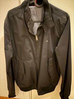 Svart vindjacka från J.Lindeberg - Stilren svart vindjacka från J.Lindeberg med diskret logga på bröstet. Jackan har hög krage, dragkedja framtill och två snedställda fickor. Tillverkad i ett lätt och smidigt material, perfekt för blåsiga dagar.