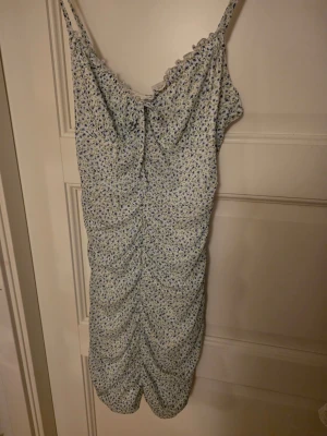 Snygg oanvänd klänning - Den är oanvänd och lappen sitter kvar. Den har ett stechigt material och lägger sig snyggt rund kroppen. Söt blommig kortklänning från H&M med smala axelband och rynkade detaljer framtill. Klänningen har en ljus bas med små blå och gröna blommor och är gjord i återvunnen polyester. Perfekt för sommarens alla tillfällen!