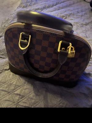 handväska brun -  handväska i Damier-mönster. Väskan är brun med mörkbruna detaljer och guldiga metalldelar. Insidan är vinröd och har ett fack. Perfekt storlek för vardagens essentials och har dragkedja upptill.