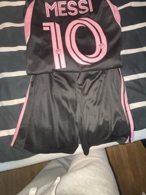 Inter Miami Messi matchställ svart/rosa - Snyggt matchställ från Inter Miami med Messi #10. Setet består av en svart tröja och shorts med rosa detaljer, klubbmärke och Adidas-logga. Tillverkat i lätt och ventilerande material, perfekt för fotbollsträning eller match.