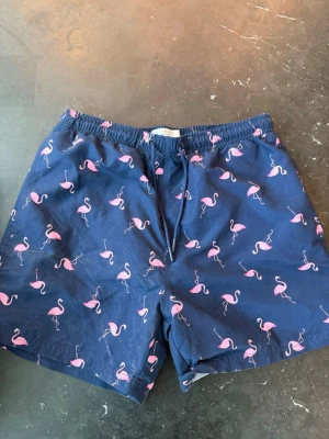 Mörkblå badbyxor med flamingos - Snygga mörkblå badbyxor med elastisk midja och snörning. Mönstrade med rosa flamingos över hela tyget för en lekfull och somrig vibe. Perfekta för stranden eller poolen. Tillverkade i snabbtorkande material.