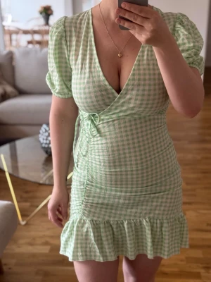 Grön- och vit rutig omlottklänning - Helt oanvänd kort omlottklänning i ljusgrönt och vitt rutigt mönster. Klänningen har puffärmar, v-ringning, knyt i midjan och volangkant nertill. Något längre baktill.  Är lite för liten för mig, därav säljer jag den.