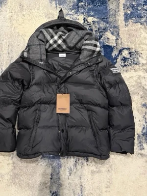 Svart pufferjacka från Burberry - Säljer en svart pufferjacka från Burberry med klassiskt rutigt foder i huvan. Jackan har dragkedja och knappar framtill, två snedställda fickor och en tydlig Burberry-patch på ärmen. Perfekt för kalla dagar och riktigt snygg till streetwear.