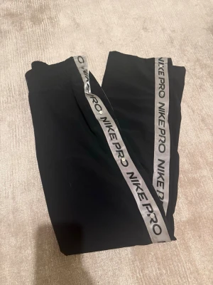 Nike Pro svarta träningsbyxor - Säljer ett par svarta Nike Pro träningsbyxor med bred resår i midjan och gråa sidoremsor med Nike Pro-logga. Byxorna är i stretchigt material och har en sportig look, perfekta för gymmet eller löpning.