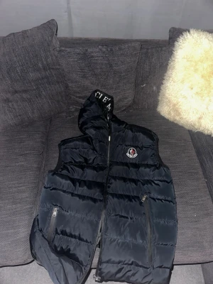 Svart dunväst från Moncler - Säljer en svart dunväst från Moncler med ikonisk logga på bröstet och stora Moncler-tryck på insidan. Västen har quiltad design, dragkedja framtill och två fickor med dragkedja. Perfekt för lager-på-lager och riktigt snygg till streetwear.