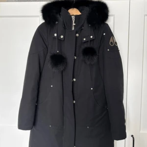 Svart parkas från Moose Knuckles - Säljer en svart parkas från Moose Knuckles med fluffig pälskantad huva och två stora pälstofsar. Jackan har dragkedja, knappar och logga på ärmen. Som ny, använd ett fåtal gånger. Perfekt för kalla dagar.