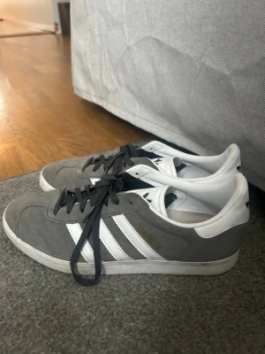 Adidas Gazelle grå/vita sneakers - Säljer ett par klassiska Adidas Gazelle sneakers i grå mocka med vita detaljer och svarta skosnören. Skorna har den ikoniska trefoil-loggan på hälen och tungan samt vita ränder på sidorna. Perfekta för dig som gillar retrostil och streetwear. Skorna är använda fåtal gånger! Och vita skosnören ingår! 