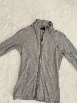 Grå tränings hoodie - Säljer en ljusgrå hoodie från Gina Tricot i storlek S. Tröjan har dragkedja framtill, hög krage och långa ärmar. Tillverkad i mjuk bomullsblandning, perfekt för chill dagar eller till träningen.