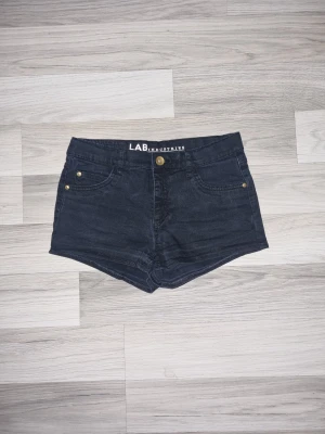 Svarta denimshorts från LAB Industries - Snygga svarta denimshorts från LAB Industries i klassisk femficksmodell med knapp och dragkedja framtill. Shortsen har guldiga nitar vid fickorna och är midwaist. Perfekta för varma dagar och har en normal passform.