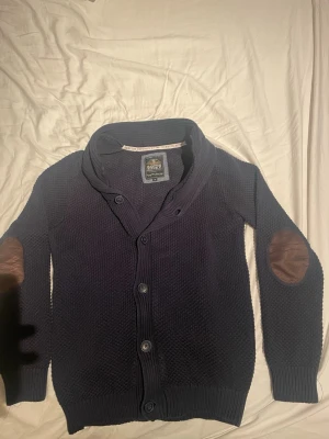 Mörkblå kofta Hampton Republic - Säljer en mörkblå stickad kofta från Hampton Republic i storlek M. Koftan har snygga bruna mockadetaljer på armbågarna, bred krage och knäppning med fyra knappar framtill. Perfekt för dig som gillar klassisk och chill stil🌟