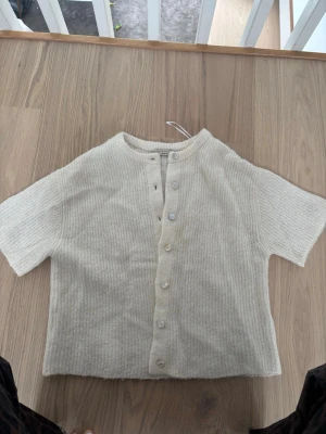 Beige ribbad kofta från Gina Tricot - Kortärmad beige kofta från Gina Tricot i mjuk stickad kvalitet. Tröjan har ribbad struktur, rund halsringning och knappar framtill. Perfekt att slänga över ett linne eller t-shirt för en chill look.