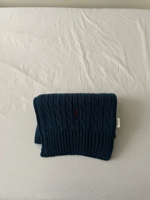 Ralph Lauren halsduk - Säljer en mörkblå kabelstickad halsduk från Polo Ralph Lauren med klassisk röd broderad logga. Halsduken är i mjuk ull och har en chunky känsla som passar perfekt till kyliga dagar. Snygg och tidlös accessoar till din vinterstil.