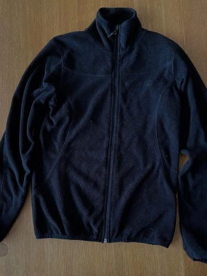 Svart fleecejacka från Etirel - Mysig svart fleecejacka från Etirel med hel dragkedja framtill och hög krage. Perfekt för kyliga dagar och enkel att slänga på över en t-shirt. Klassisk och enkel design som funkar till det mesta.