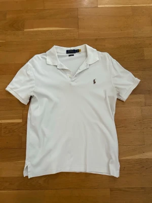 Vit pikétröja Polo Ralph Lauren  - Klassisk vit pikétröja från Polo Ralph Lauren i slim fit-modell. Tröjan har korta ärmar, krage och två knappar vid halsen. Ikoniska broderade loggan på bröstet. Perfekt till jeans eller shorts för en clean och stilren look.
