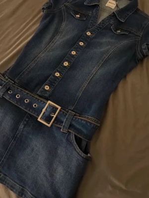 Jeansklänning med bälte från Bershka - Säljer en snygg jeansklänning från Bershka i mörkblå denim. Klänningen har korta ärmar, knappar hela vägen framtill och ett matchande bälte i midjan. Fickor framtill och klassisk skjortkrage ger en cool vibe. Perfekt för dig som gillar denim!
