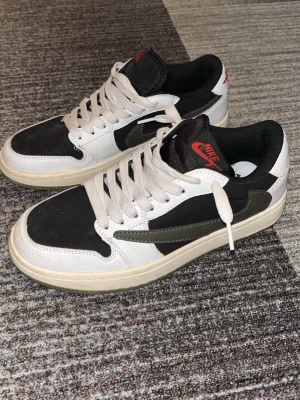 Nike Air Jordan 1 Low Travis Scott - Säljer ett par Nike Air Jordan 1 Low Travis Scott med svart och vit ovandel i skinn och mocka, olivgröna detaljer och omvänd swoosh. Röda broderade loggor på hälen och tungan samt 'Cactus Jack'-text på sidan. Klassisk platt sula och snörning.