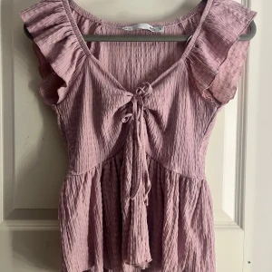 Ljusrosa blus med volang från 157 - Supersöt ljuslila blus från 157 med volangärmar och knyt framtill. Blusen har en lätt rynkad struktur och är tillverkad i ett mjukt, tunt tyg som ger en luftig känsla. Perfekt till sommaren och har en feminin, flowy passform.