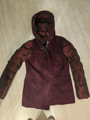 Vinröd hybridjacka från Tommy Hilfiger - Säljer en vinröd jacka från Tommy Hilfiger i storlek S. Jackan har quiltade, glansiga ärmar och rygg i pufferstil, medan framstycket och huvan är i ullmix. Fyra stora fickor framtill, dragkedja och tryckknappar. Perfekt för höst och vinter! 