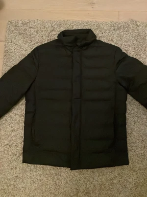 Sv rising pufferjacka från Massimo Dutti - Säljer en svart pufferjacka från Massimo Dutti med hög krage och diskret dragkedja framtill. Jackan har en stilren look med horisontella sömmar och är perfekt för kyliga dagar. Tillverkad i syntetmaterial och har långa ärmar.