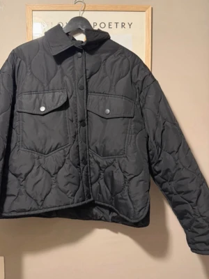 Svart quiltad jacka från Amisu - Quiltad svart jacka från Amisu i storlek 40. Jackan har en loose passform, stora fickor med lock och tryckknappar framtill. Tillverkad i polyester och har långärmade ärmar. Perfekt för höst och vinter när du vill ha en chill och trendig look. Litet hål i ena armbågen, se bild. Går att lag med lim, eller sy