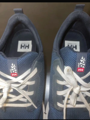 Blå sneakers från Helly Hansen - Säljer ett par blå sneakers från Helly Hansen med vit sula och vita snören. Ovandelen är i mesh för bra andningsförmåga och har HH-logga på sidan. Skorna har rund tå och klassisk snörning. Perfekta för dig som gillar sportig och clean stil. Originalpris: 1200kr 