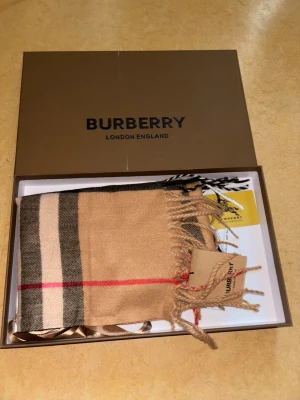 Rutig beige halsduk från Burberry - Säljer en klassisk rutig halsduk från Burberry i beige, svart, vitt och rött. Halsduken är tillverkad i 100% kashmir och har fransar i ändarna. Den ikoniska rutiga designen gör den superenkel att styla till olika outfits. Materialet är mjukt och skönt.
