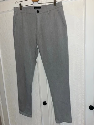 Grå kostymbyxor från Tailored Originals - Snygga grå kostymbyxor från Tailored Originals med klassisk rak passform. Byxorna har bälteshällor, sidofickor och bakfickor med knapp. Materialet är mjukt och har en lätt melerad look. Perfekta till skjorta eller stickad tröja. 