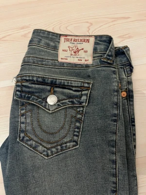 True Religion Joey Low Rise Flare Jeans - Säljer dessa äkta True Religion jeans i klassisk blå tvätt med den ikoniska bakfickesömmen. Väldigt trendiga och svåra att få tag på i bra skick  De är endast använda ca 5 gånger och i mycket fint skick. Säljs endast för att de tyvärr är för stora i midjan för mig. Perfekta för en vintage/Y2K streetstyle-look. TIPS: dem töjer sig mycket i midjan när man har på sig dem, köp mindre storlek. Pris kan diskuteras 